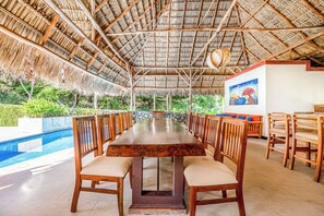 A long dining table with chairs, a pool, and a thatched roof structure.