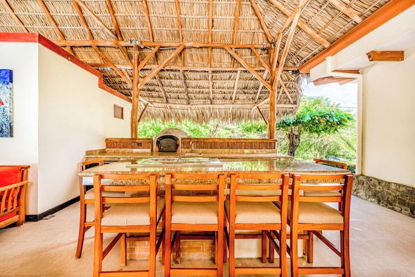 A covered outdoor dining area with a thatched roof, a stone counter, and wooden chairs. - Private Pool, Luxe Patio: 4BR Carrillo Villa (Sardinal)
