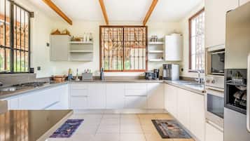 Una cocina moderna con armarios blancos, horno empotrado, cafetera y ventana con celosía decorativa.