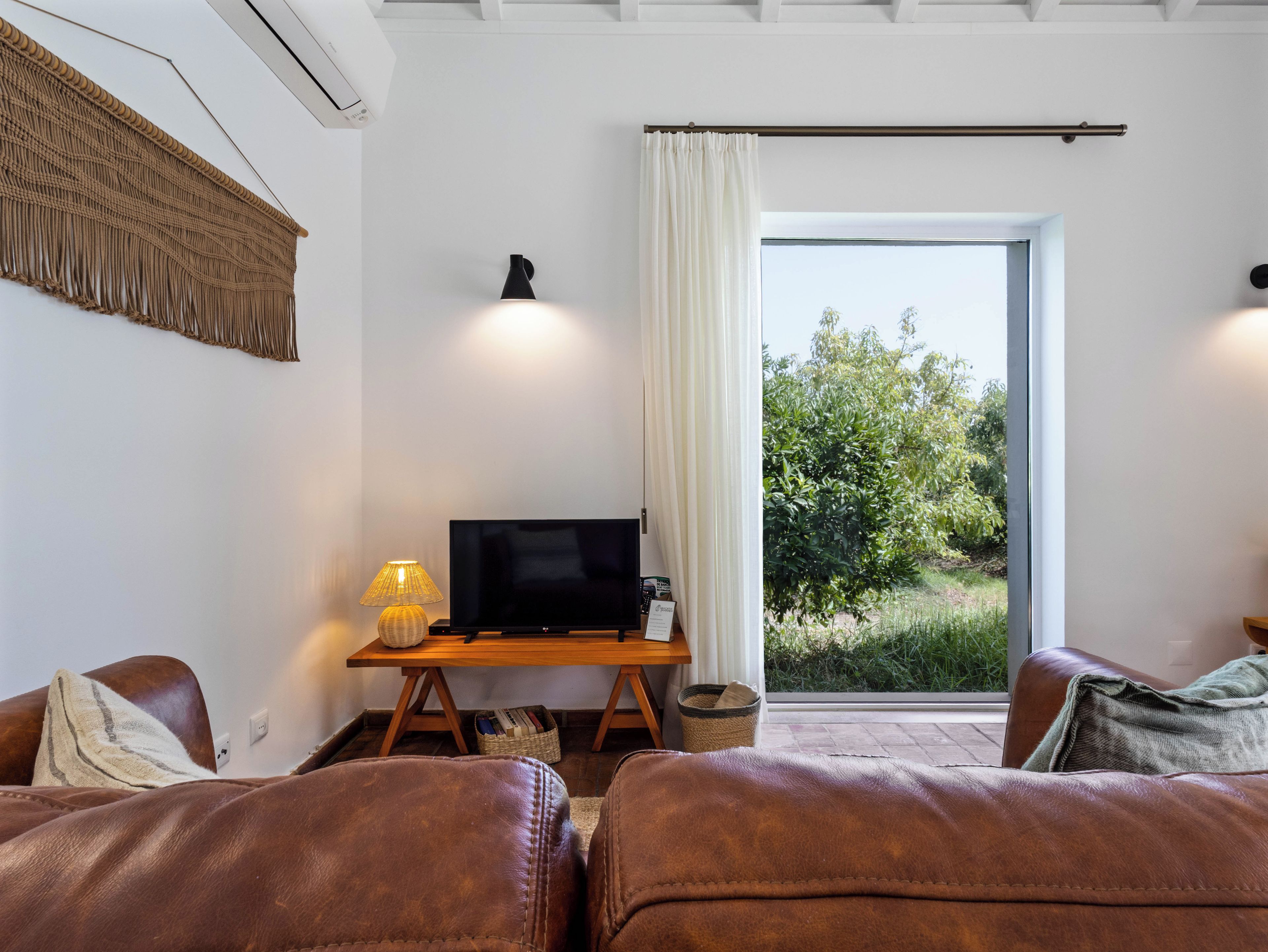 Een kamer met een televisie op een houten standaard, een lamp, een raam met gordijnen, en twee leren banken.