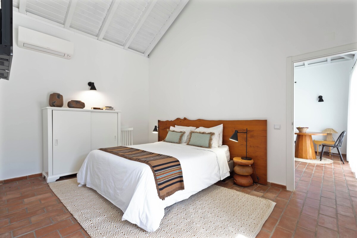 A bedroom with a large bed, a wooden headboard, a white wardrobe, and a small table with a lamp.