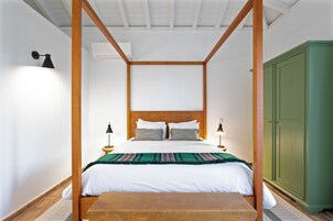 Uma cama com dossel com roupa de cama branca e várias almofadas, piso de madeira, guarda-roupa verde e uma pequena mesa de madeira.