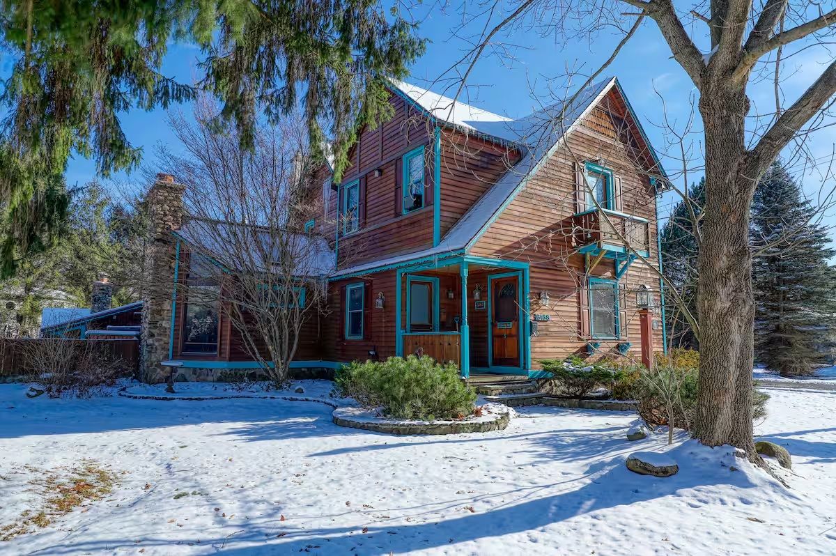 A two-story wooden house with a snow-covered roof, surrounded by trees and a snow-covered ground.