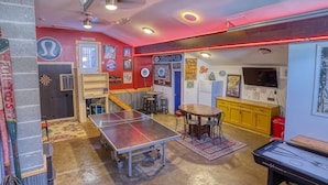 A room with a ping pong table, a dartboard, and a pool table. There are stools and a table with chairs. The walls have various posters and signs. - Large Ski Home: Seasonal Pool & Year-round Hot Tub (Ellicottville)