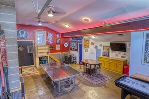 Um quarto com mesa de pingue-pongue, dardos e mesa de bilhar. Há banquetas e uma mesa com cadeiras. As paredes possuem diversos cartazes e placas.