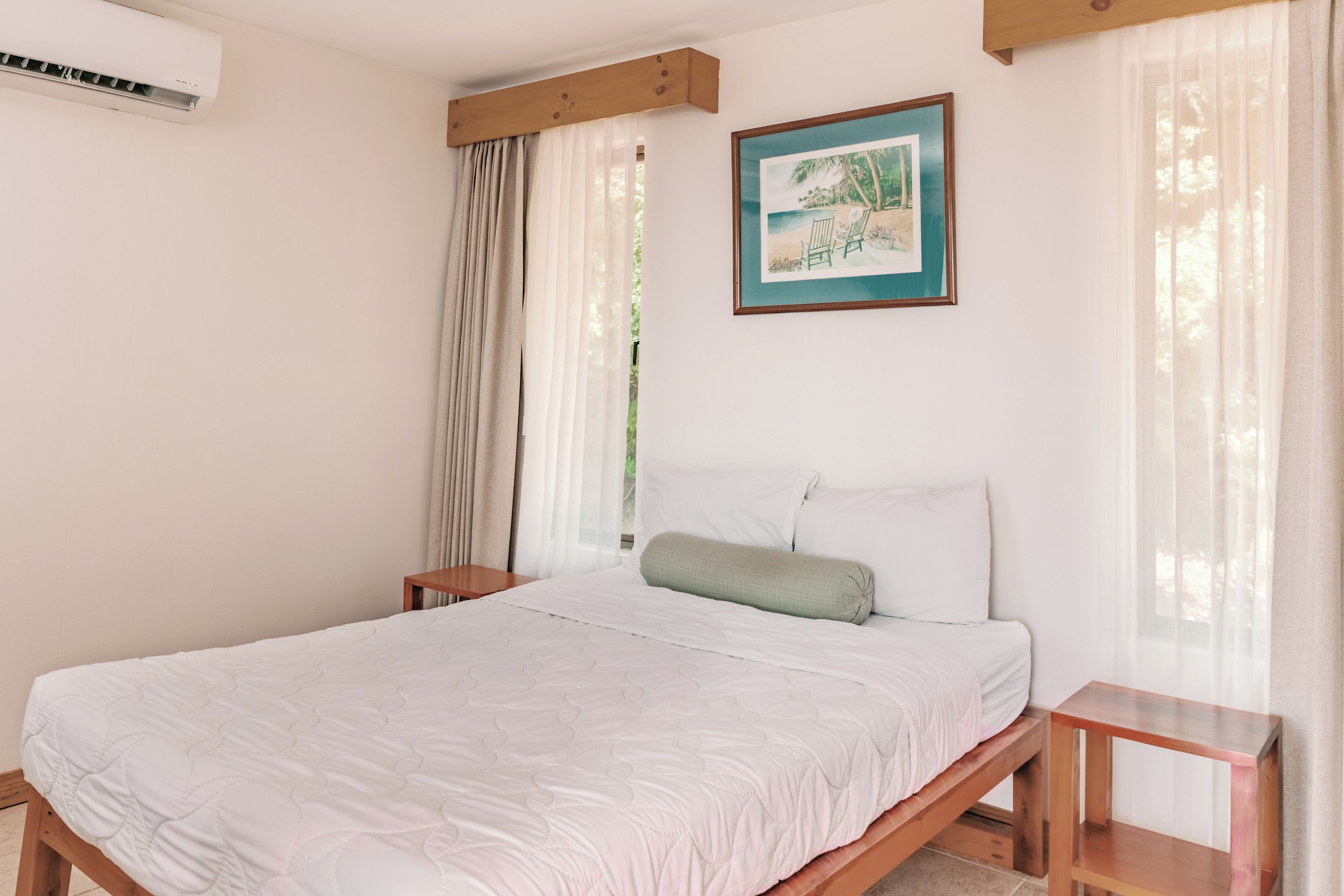Un dormitorio con cama, mesita de noche, cabecero de madera, un cuadro en la pared y ventana con cortinas traslúcidas.