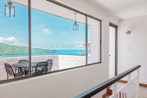 Varanda com mesa e cadeiras, com vista para a paisagem costeira com colinas e o mar.