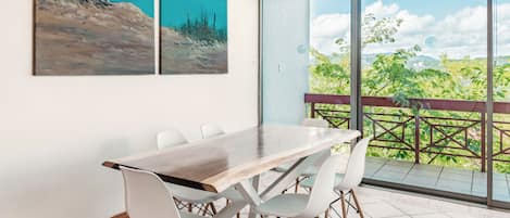 A dining area with a table and chairs, large windows with a view of greenery and a building, and two paintings on the wall.