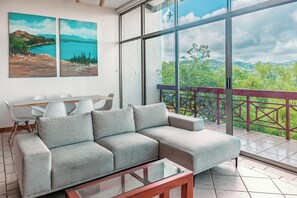 A modern living room with a sectional sofa, a glass coffee table, and a dining area with a white table and chairs. There is a large window with a view of greenery and a distant landscape. - Playa del Coco 3br: Ocean Views & Pools! (Sardinal)