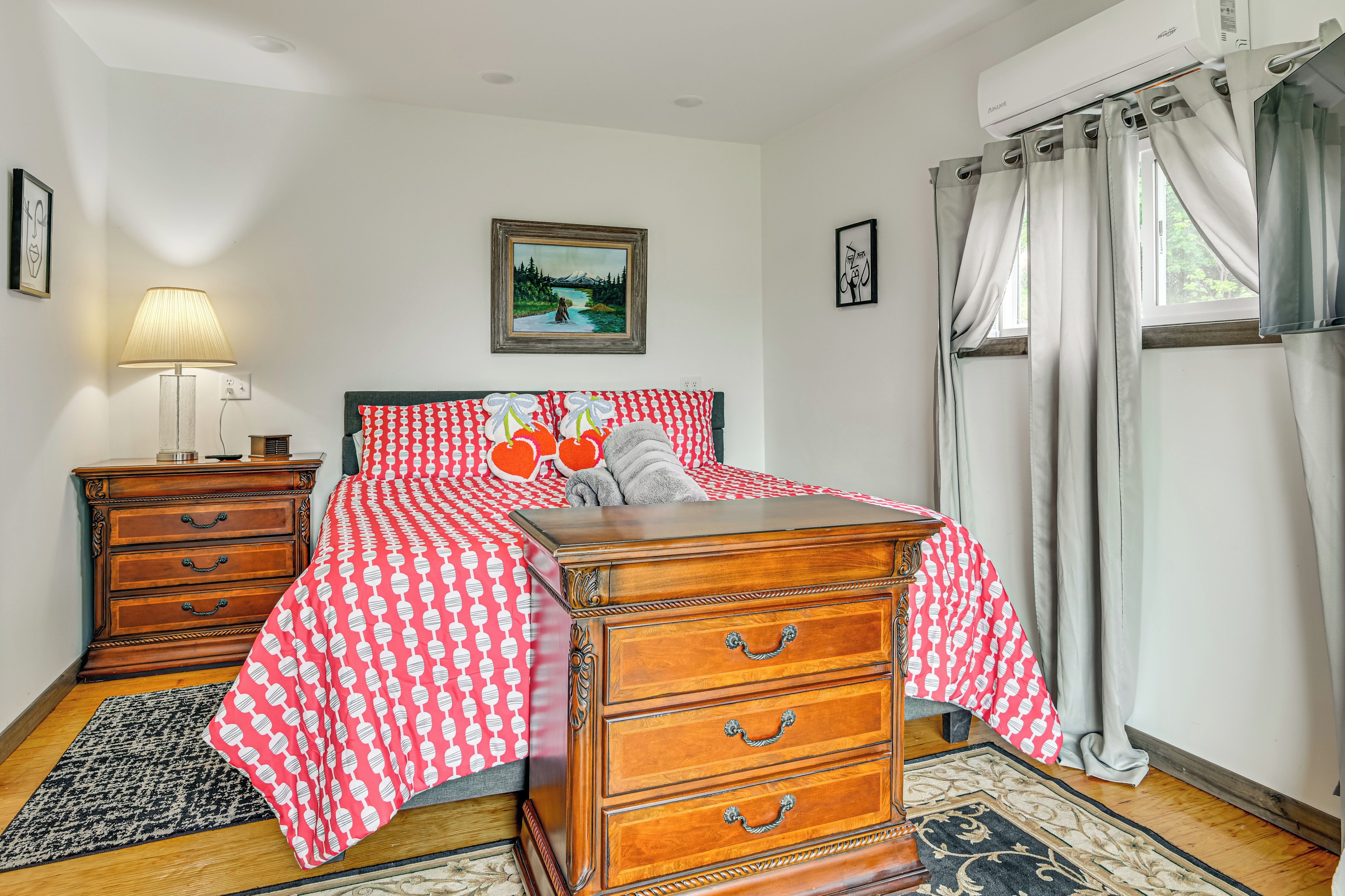 A bedroom with a wooden dresser, a bed with a red and white patterned bedspread, a nightstand with a lamp, and a painting on the wall.