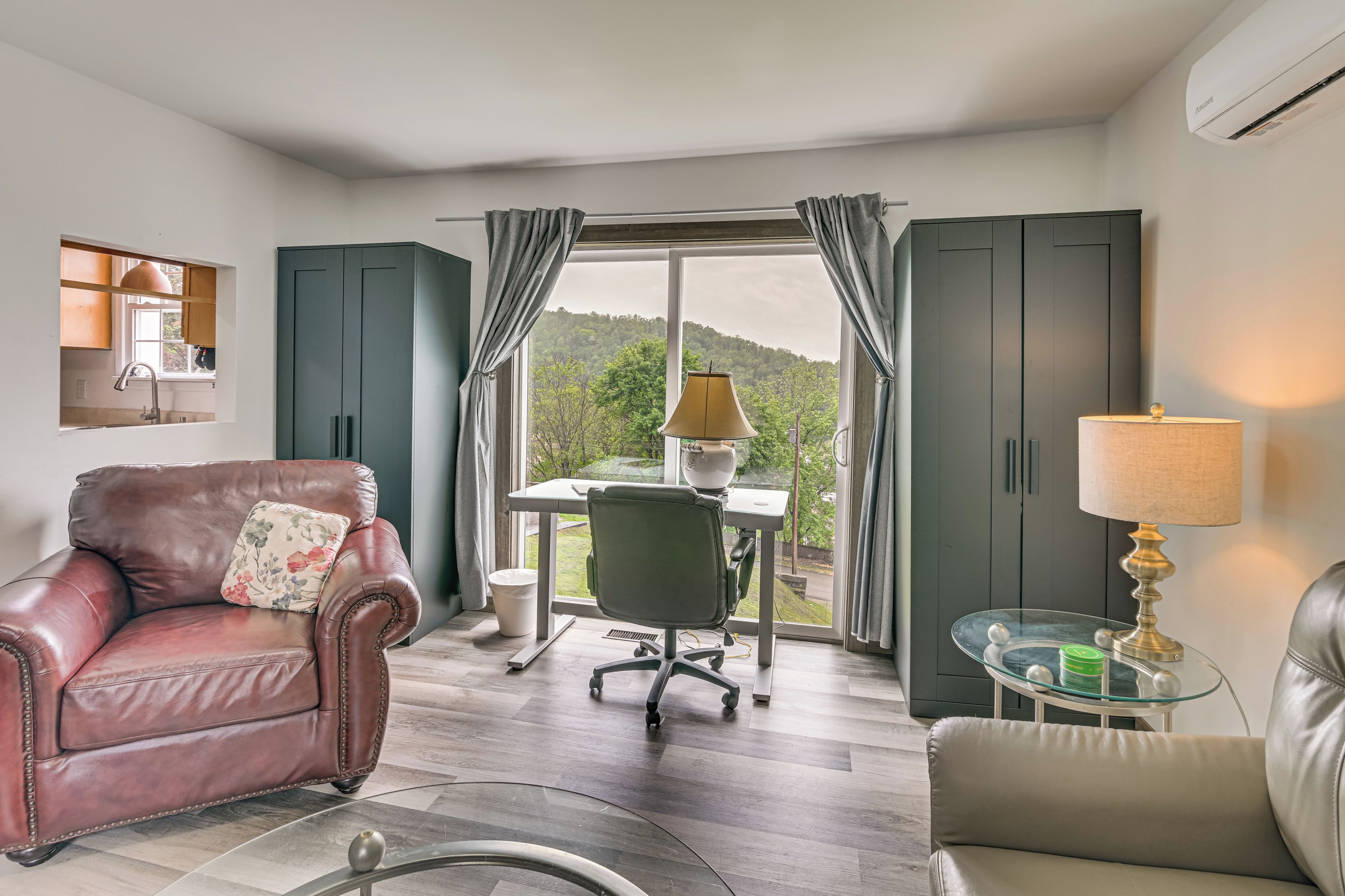 A modern living room with a leather sofa, a desk with a chair, a glass coffee table, and a view of a green landscape through large windows.