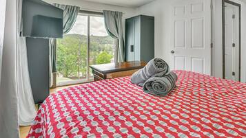 A hotel room with a large bed, a wooden nightstand, a desk, and a view of greenery through the window.