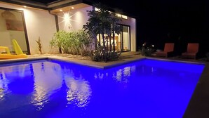 Una piscina con bordo illuminato di blu, circondata da una casa con una porta in vetro e piante in vaso.
