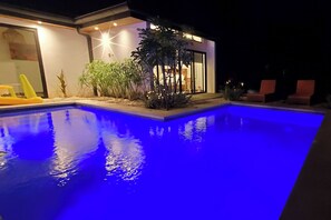 A swimming pool with a blue-lit edge, surrounded by a house with a glass door and potted plants.