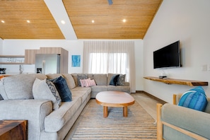 A modern living room with a sectional sofa, a round coffee table, a flat-screen TV mounted on the wall, and a wooden ceiling.