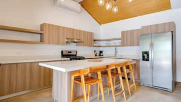 A modern kitchen with a central island, wooden bar stools, a stainless steel refrigerator, and a wooden ceiling.