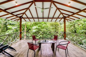 Un espacio de estar al aire libre con techo, con una mesa y sillas, rodeado de abundante vegetación.