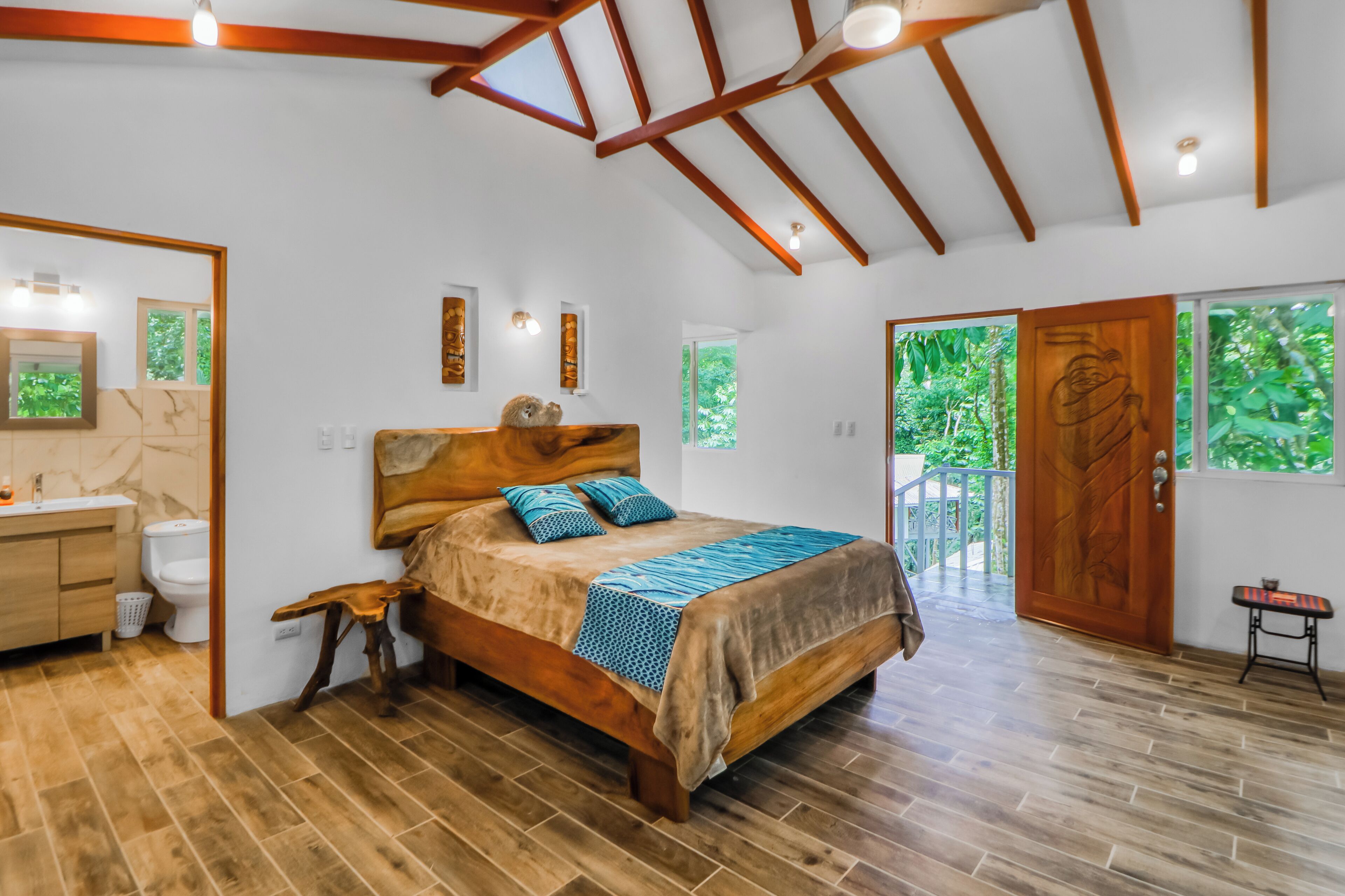 A bedroom with a wooden bed, a wooden bench, a small table, a mirror, and a view of the outdoors.