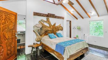 A bedroom with a large wooden headboard, a bed with patterned bedding, a wooden side table, and a view of greenery outside.
