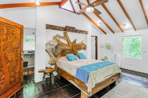 Un dormitorio con un gran cabecero de madera, una cama con ropa de cama estampada, una mesita de noche de madera y vistas a la vegetación exterior.