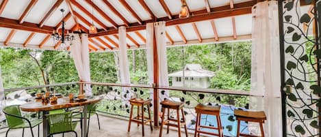 A covered outdoor area with a wooden table and chairs, a chandelier, and a view of greenery.