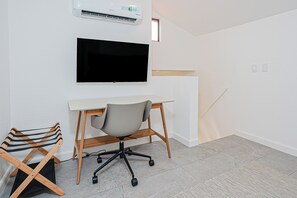 Un espace de bureau moderne et minimaliste, avec un téléviseur à écran plat fixé au mur, un bureau blanc, une chaise de bureau grise et une chaise en bois avec un coussin à rayures.