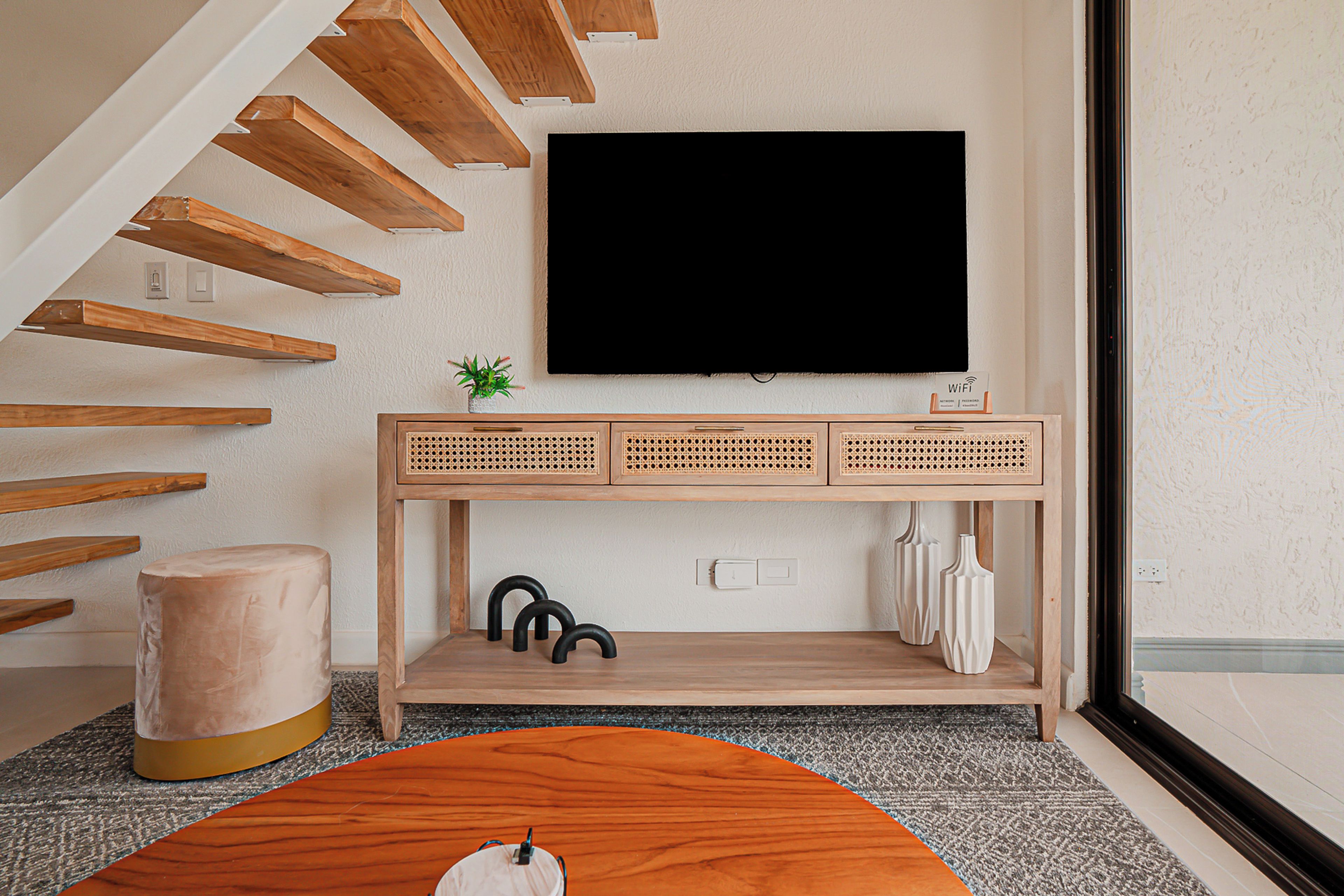 En moderne stue med en trætrappe, et fladskærms-tv, et rattan-konsolbord og en rund fodskammel.