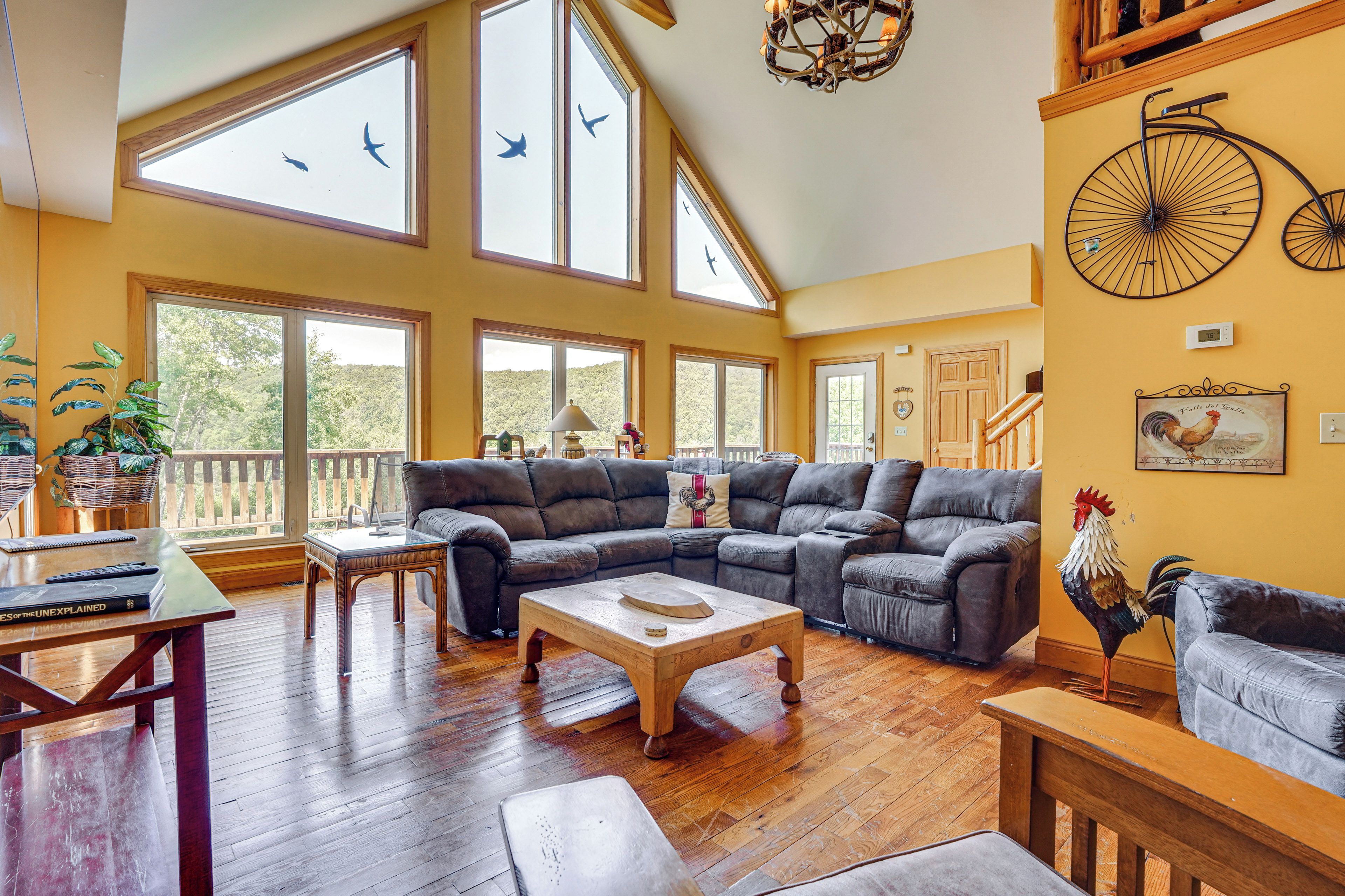 A spacious living room with a large sectional sofa, wooden flooring, and multiple windows offering views of the outdoors.