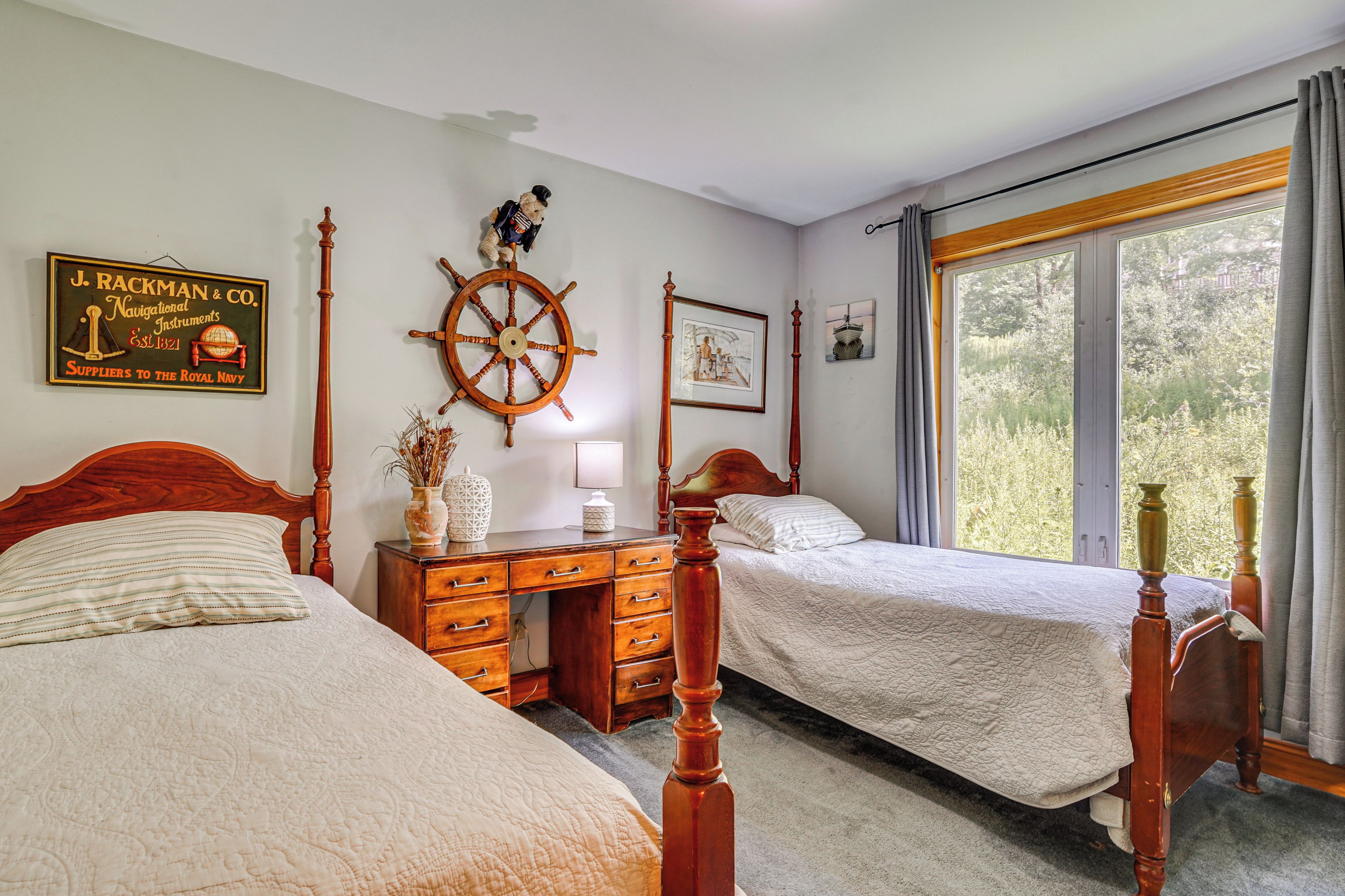 A bedroom with two wooden beds, a wooden dresser, a ship's wheel, and a window with a view of trees.