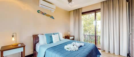 A hotel room with a bed, a ceiling fan, an air conditioning unit, a nightstand with a lamp, and a window with curtains.