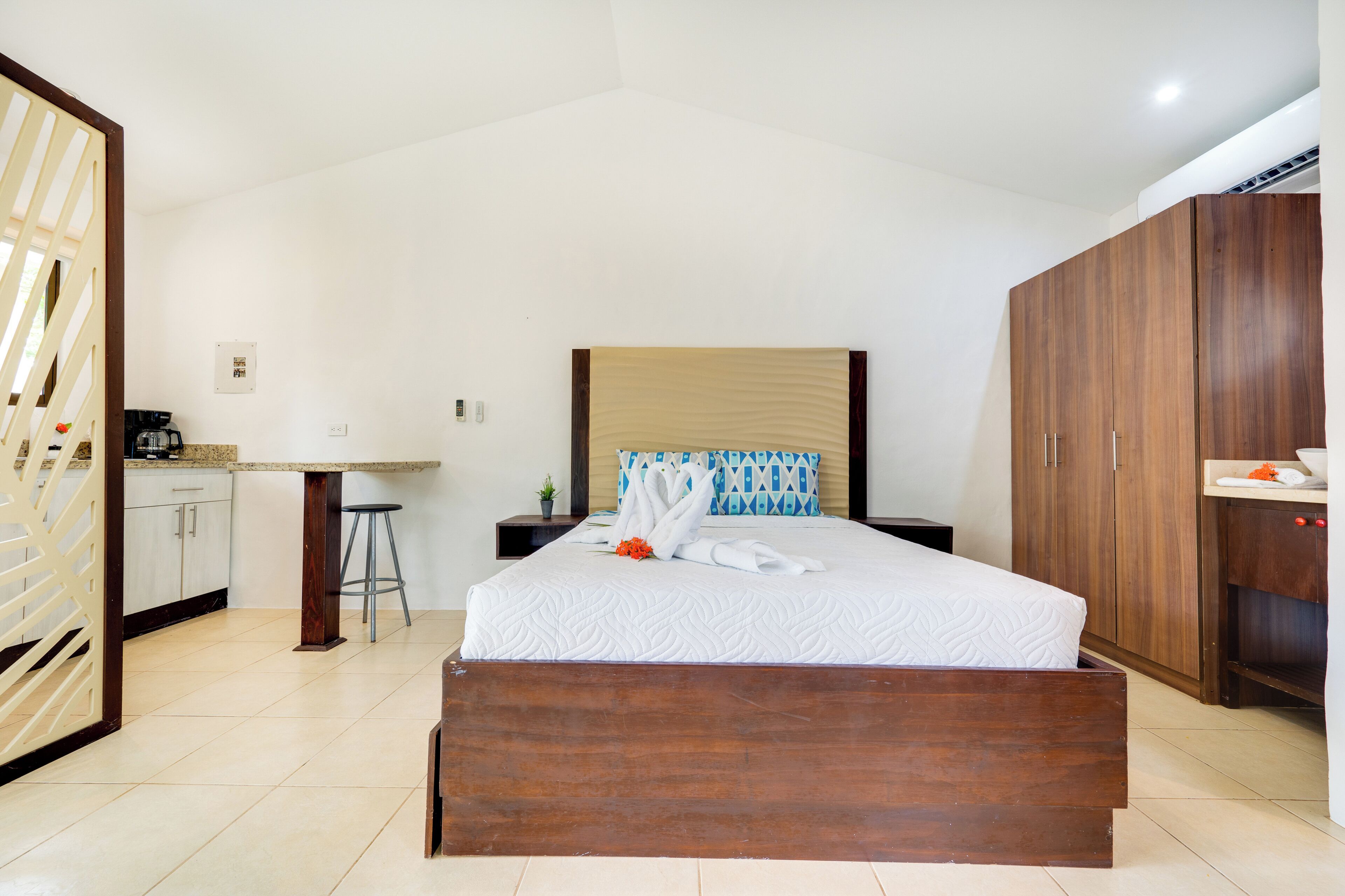 A hotel room with a bed, wooden wardrobe, a desk with a chair, and a kitchenette.