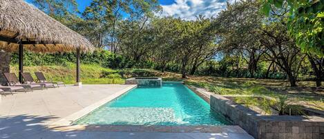 A swimming pool with a thatched roof structure, surrounded by lush greenery and trees.