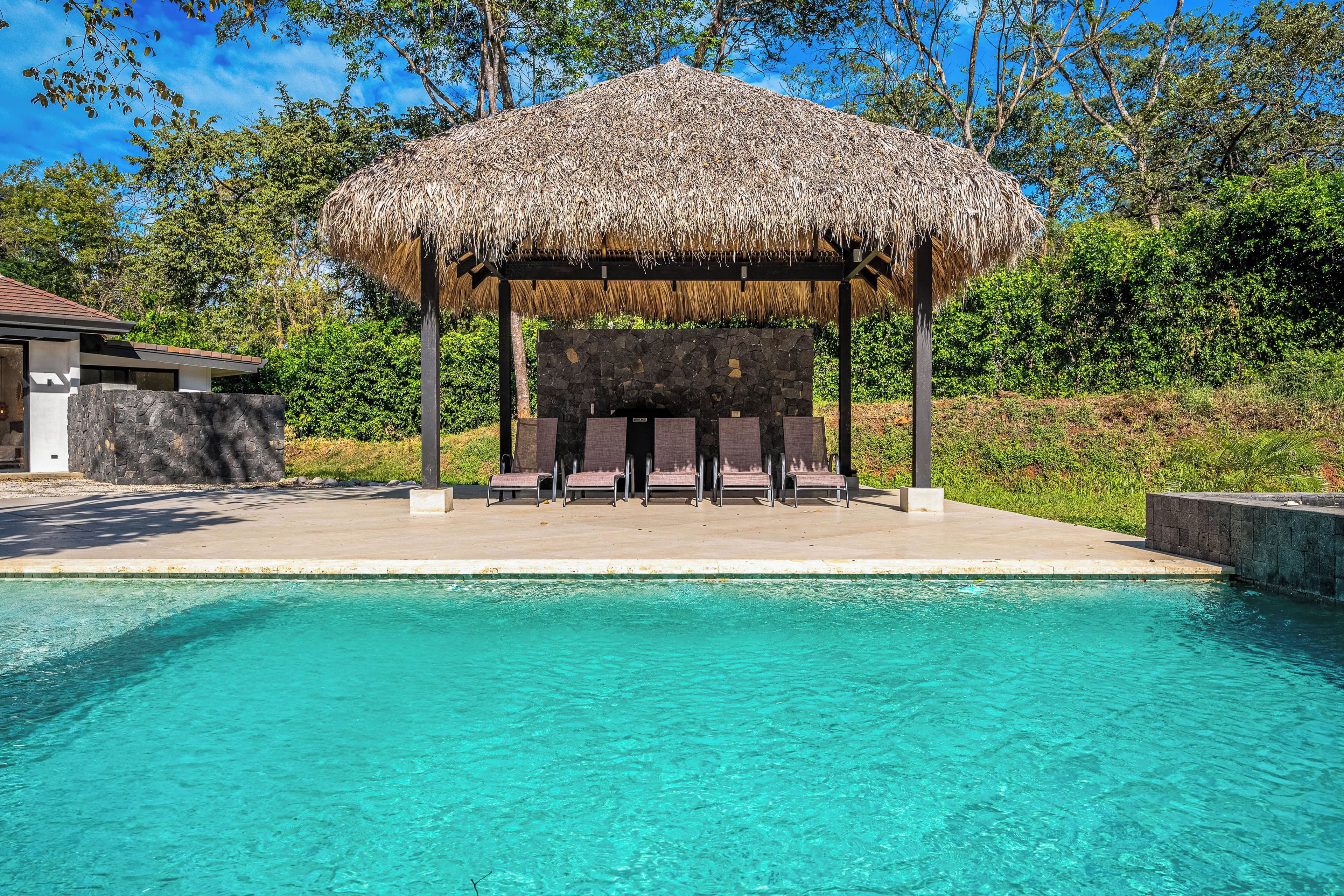 A pool area with a thatched gazebo and lounge chairs.