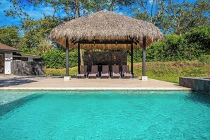 A pool area with a thatched gazebo and lounge chairs.