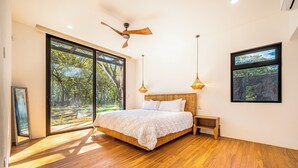 Un dormitorio con una cama grande, piso de madera, ventilador de techo y amplias ventanas que dan a vistas de árboles.