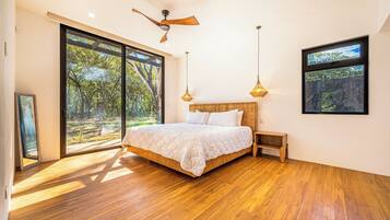 A bedroom with a large bed, wooden flooring, a ceiling fan, and large windows offering a view of trees.