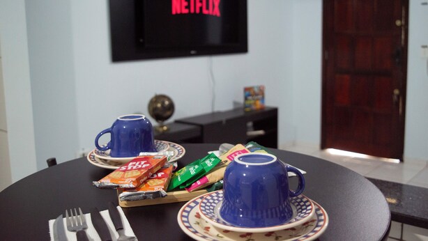A dining area with a table set for two, a television displaying the Netflix logo, and a door in the background.