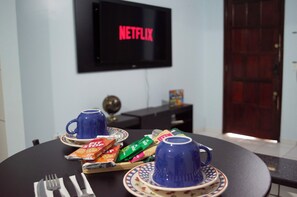 Un coin repas avec une table dressée pour deux personnes, un téléviseur affichant le logo de Netflix et une porte visible au fond.