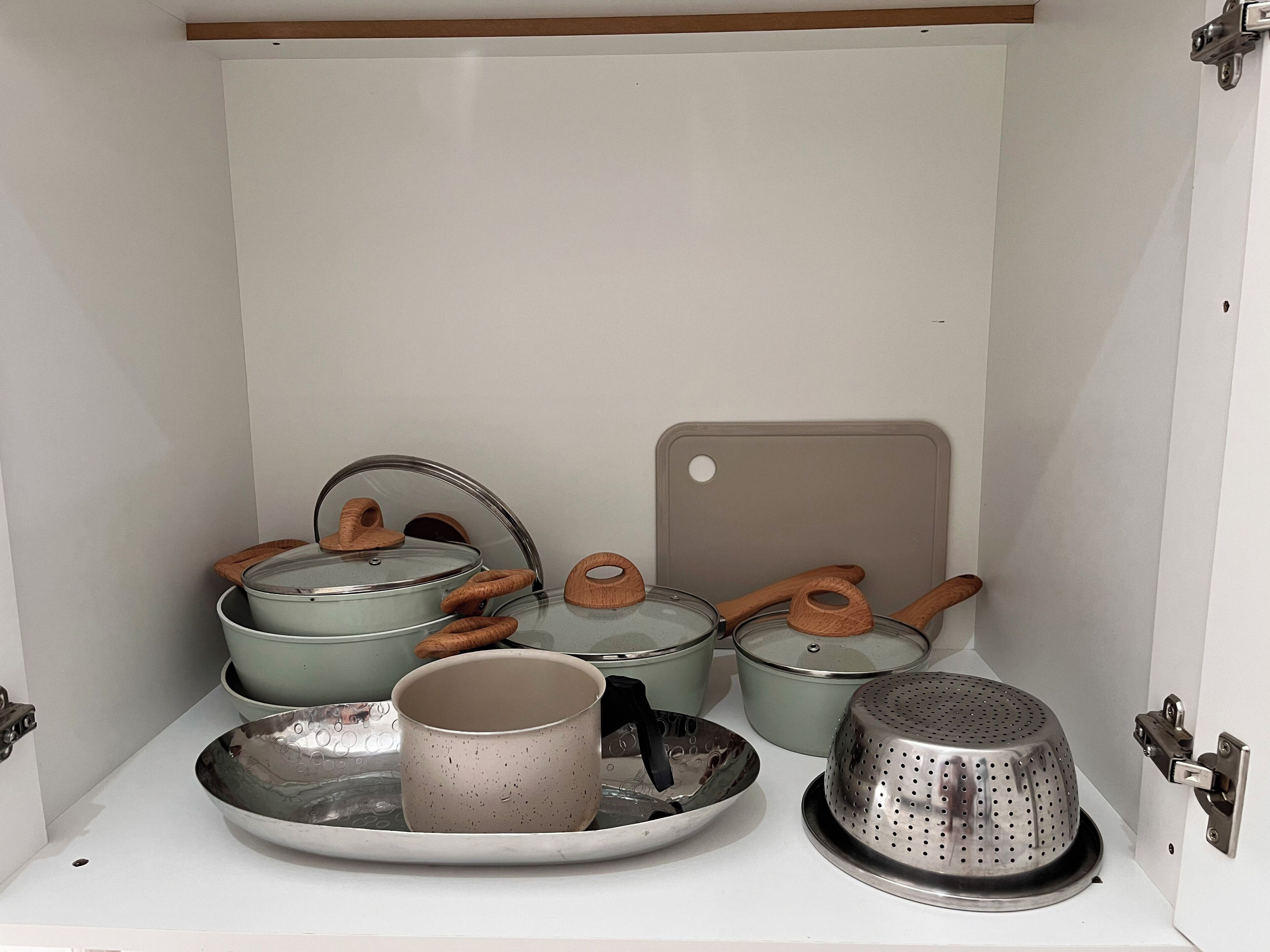 A kitchen cabinet with pots, pans, a colander, and a cutting board.