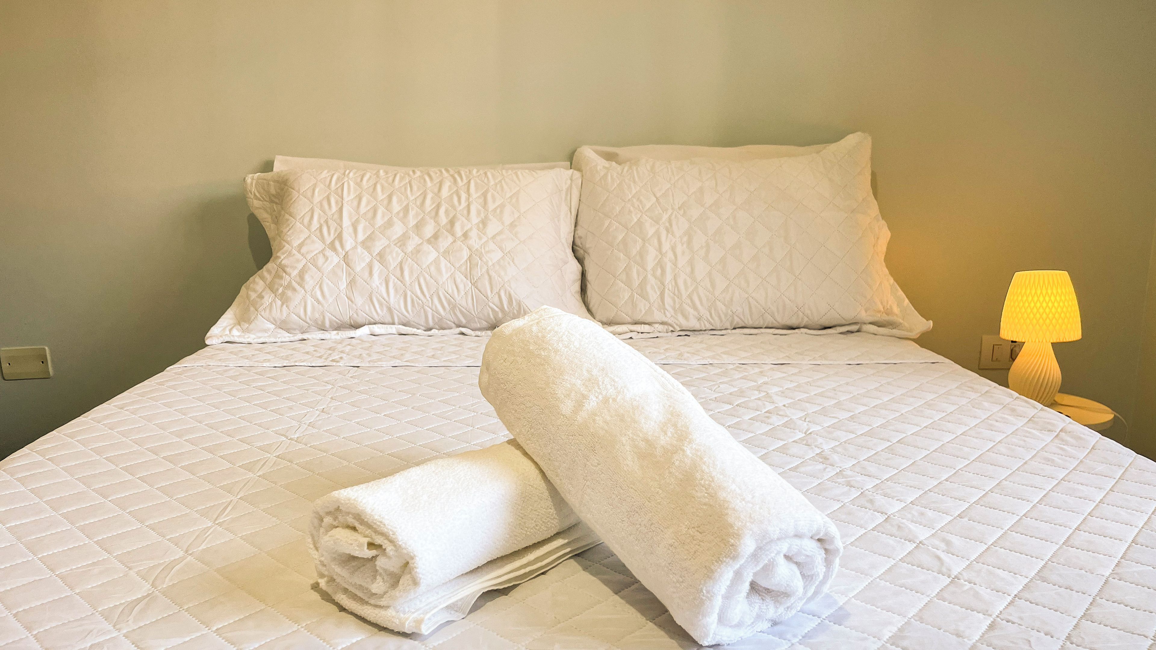 A neatly made bed with a quilted cover, two pillows, and a rolled towel on the bed.
