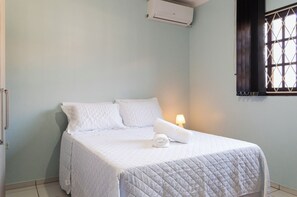Un lit simple avec une couette et des oreillers blancs matelassés, une serviette blanche enroulée et un climatiseur fixé au mur.