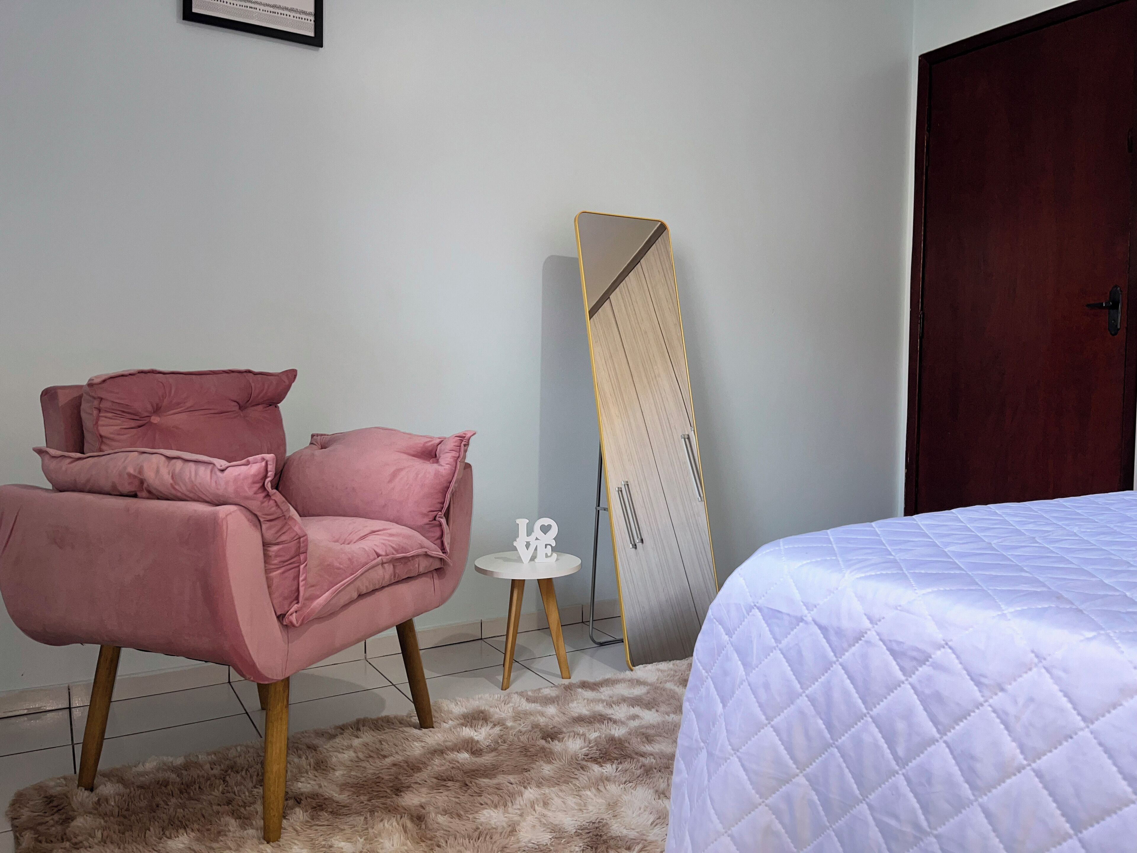 Un dormitorio con una cama, una silla rosa, una mesita blanca pequeña y un armario.