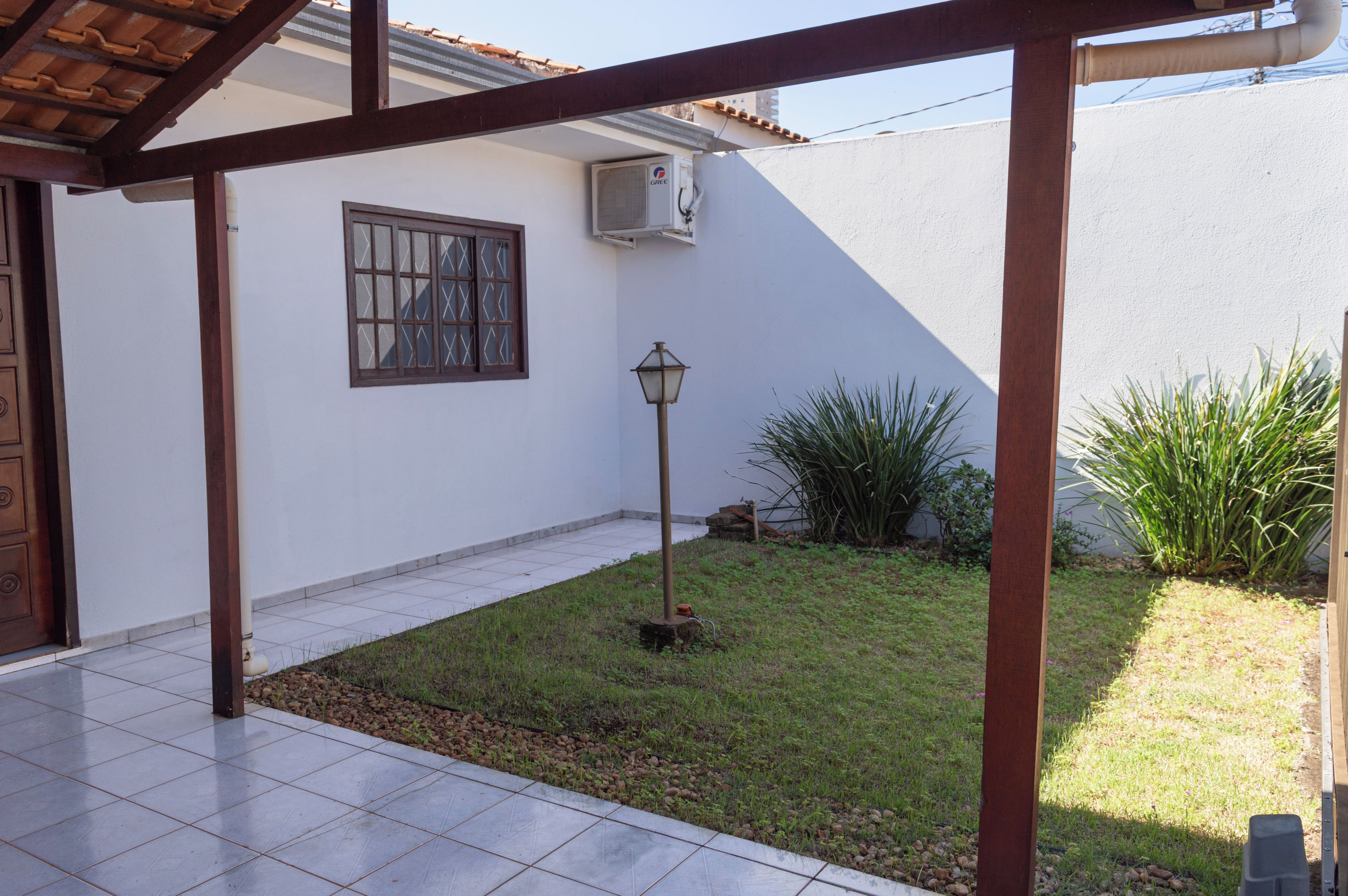 Área de varanda coberta com piso em azulejo, janela com grade de metal, ar-condicionado fixado na parede e um pequeno jardim com grama e plantas.
