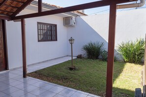 A covered patio area with a tiled floor, a window with a metal grill, a wall-mounted air conditioning unit, and a small garden with grass and plants.