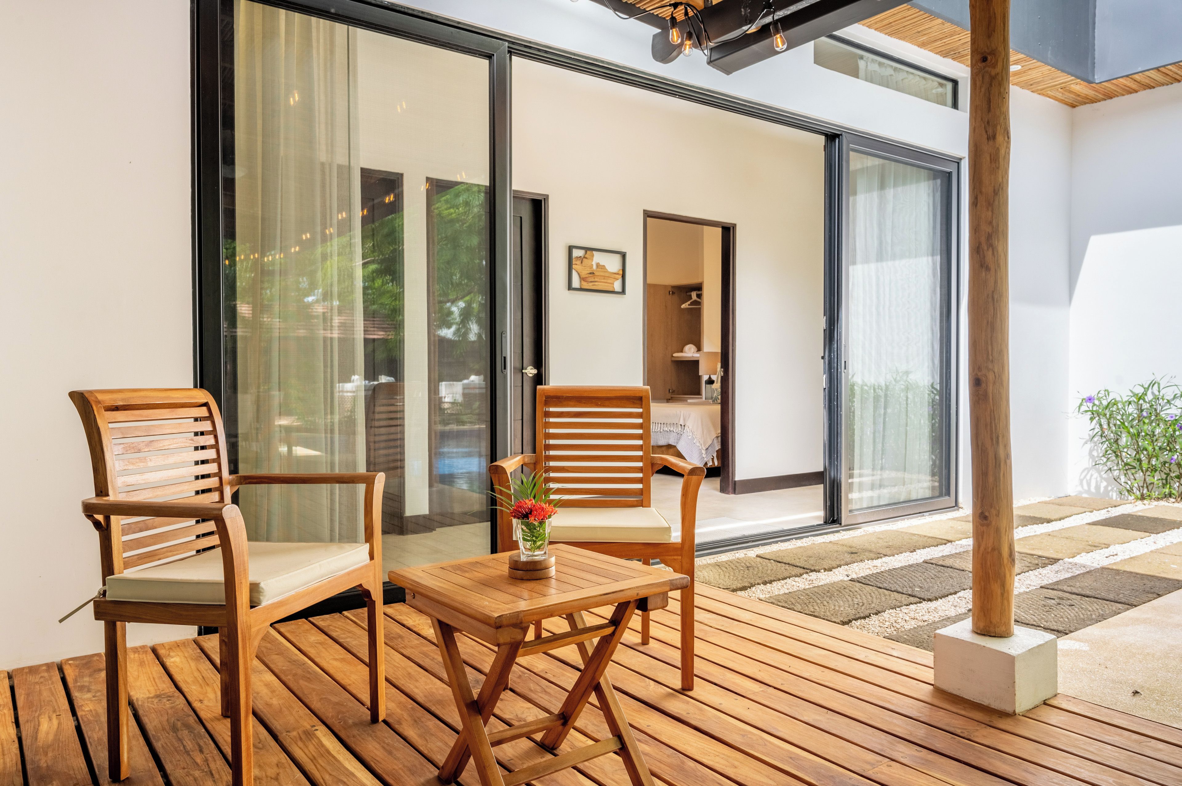 A wooden deck with two chairs and a table, a glass door leading to a room, and a wooden ceiling structure.