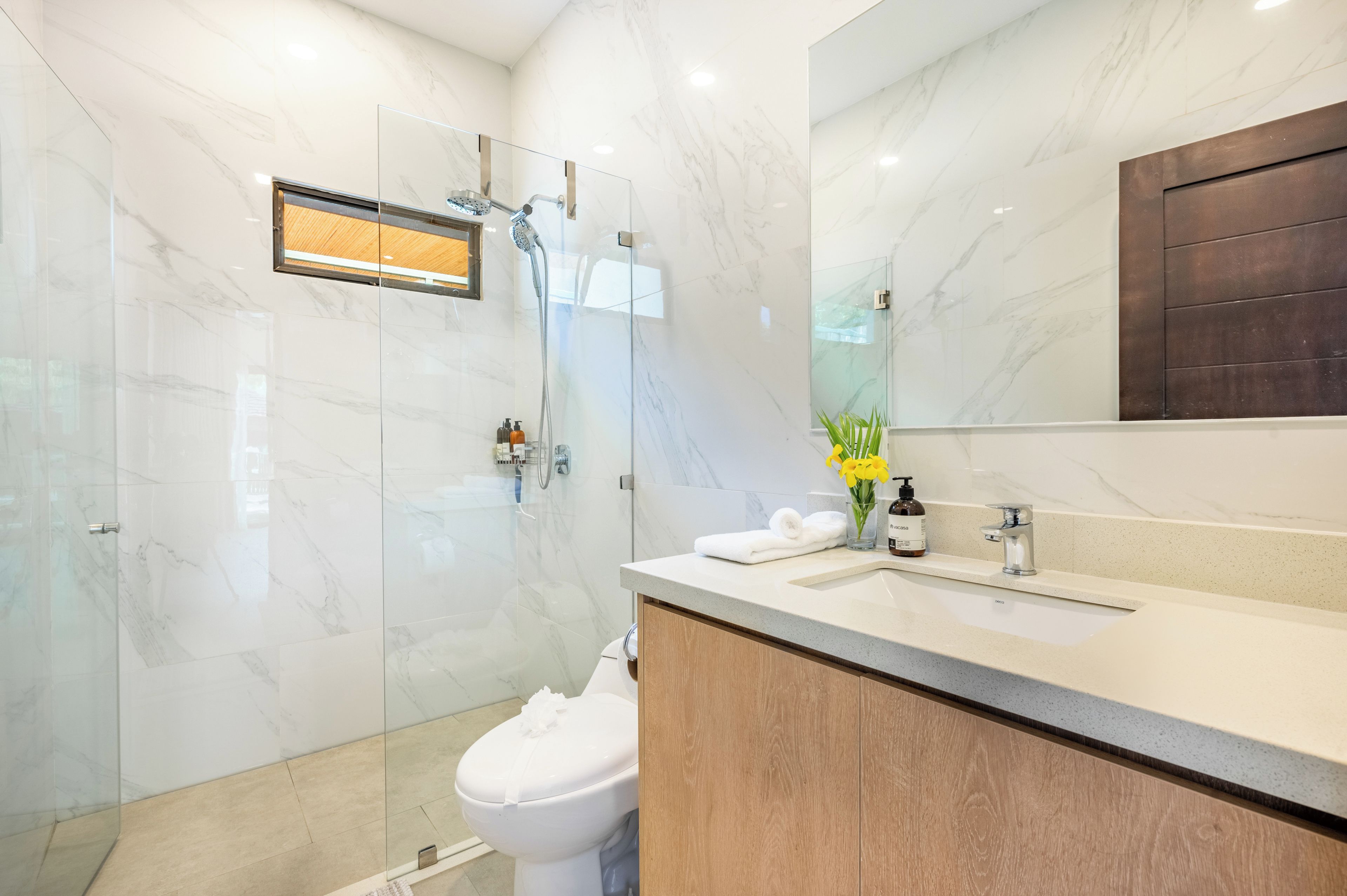A modern bathroom with a glass shower enclosure, a wooden vanity, and a marble-tiled wall.