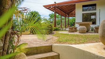 A covered outdoor area with steps leading to a building, surrounded by tropical plants and a stone bench.