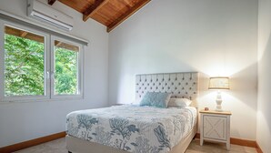A bedroom with a bed, a nightstand, a lamp, and a window with a view of trees. - Luxe Ocean-view Hideaway in Carrillo (Puerto Carrillo)