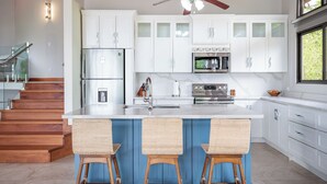 A modern kitchen with white cabinets, a central island with bar stools, stainless steel appliances, and a ceiling fan. - Luxe Ocean-view Hideaway in Carrillo (Puerto Carrillo)