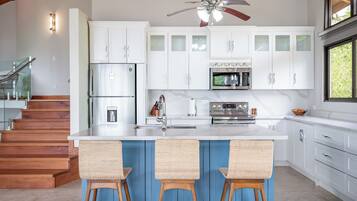 A modern kitchen with white cabinets, a central island with bar stools, stainless steel appliances, and a ceiling fan.